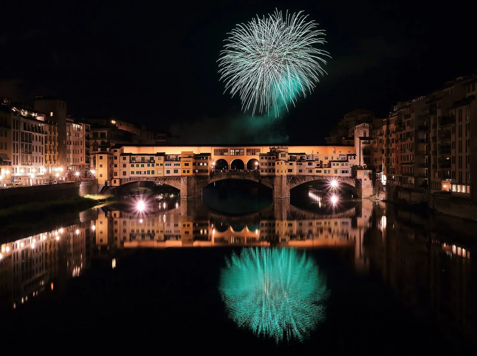 Fuochi d'artificio verdi esplodono nel cielo notturno sopra il Ponte Vecchio illuminato, con le case affacciate sull'Arno e il riflesso dei fuochi e delle luci nell'acqua del fiume