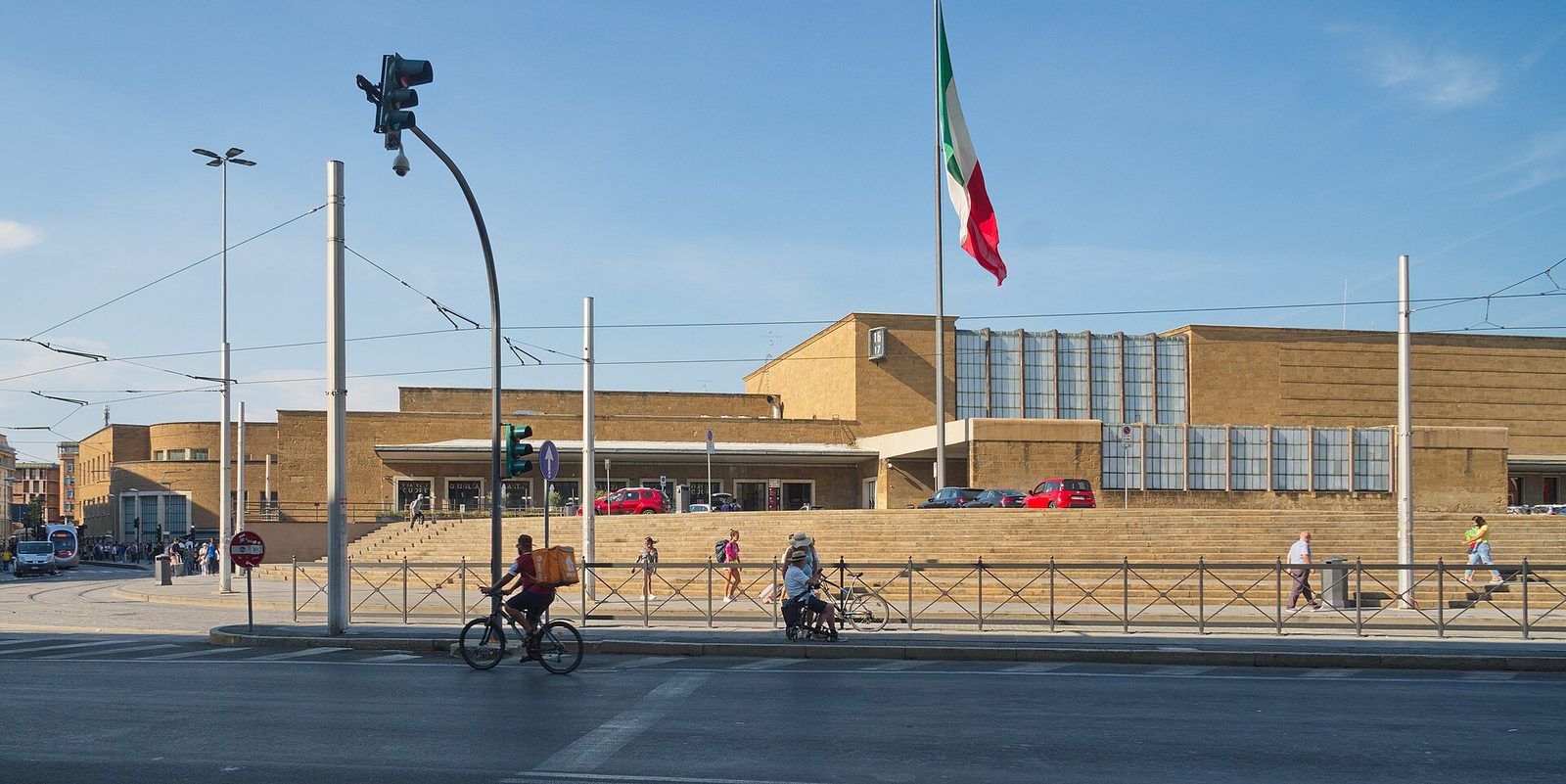 La facciata in pietra della stazione di Santa Maria Novella a Firenze, con la grande scalinata, il tricolore su un alto pennone, i binari e i pali della tramvia in primo piano e un ciclista che attraversa la strada