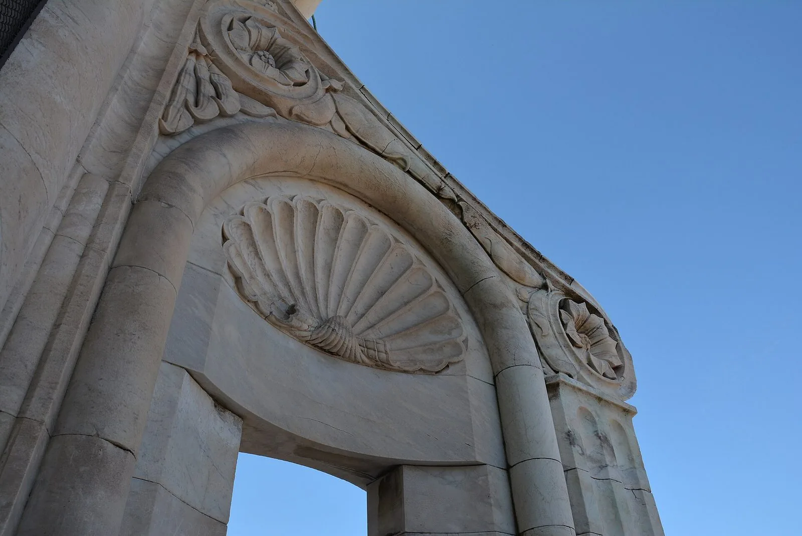 Dettaglio in marmo bianco sulla sommità della Cupola del Brunelleschi: un arco con una grande conchiglia scolpita e due rosoni a forma di fiore, visto dal basso contro un cielo azzurro intenso