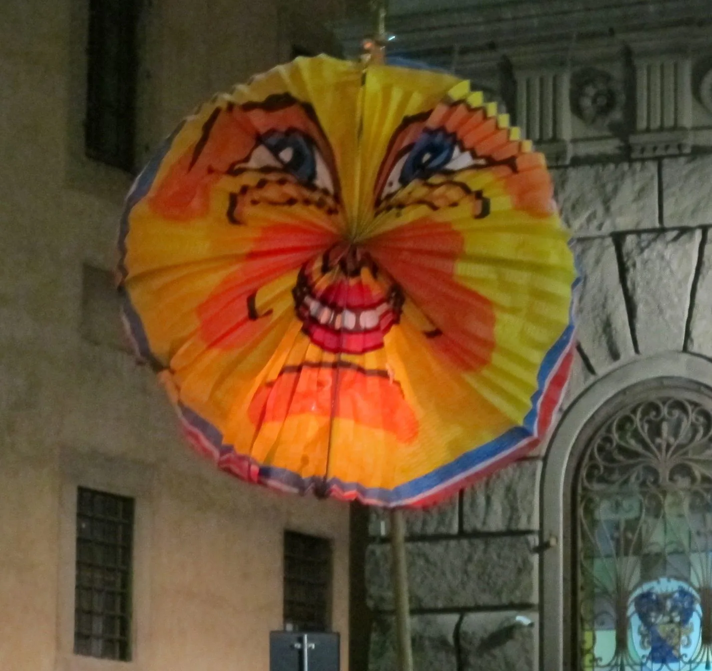 Una grande lanterna di carta tonda, gialla e arancione, dipinta con un volto di sole sorridente, sorretta da un bastone di notte davanti alla facciata in pietra di un palazzo