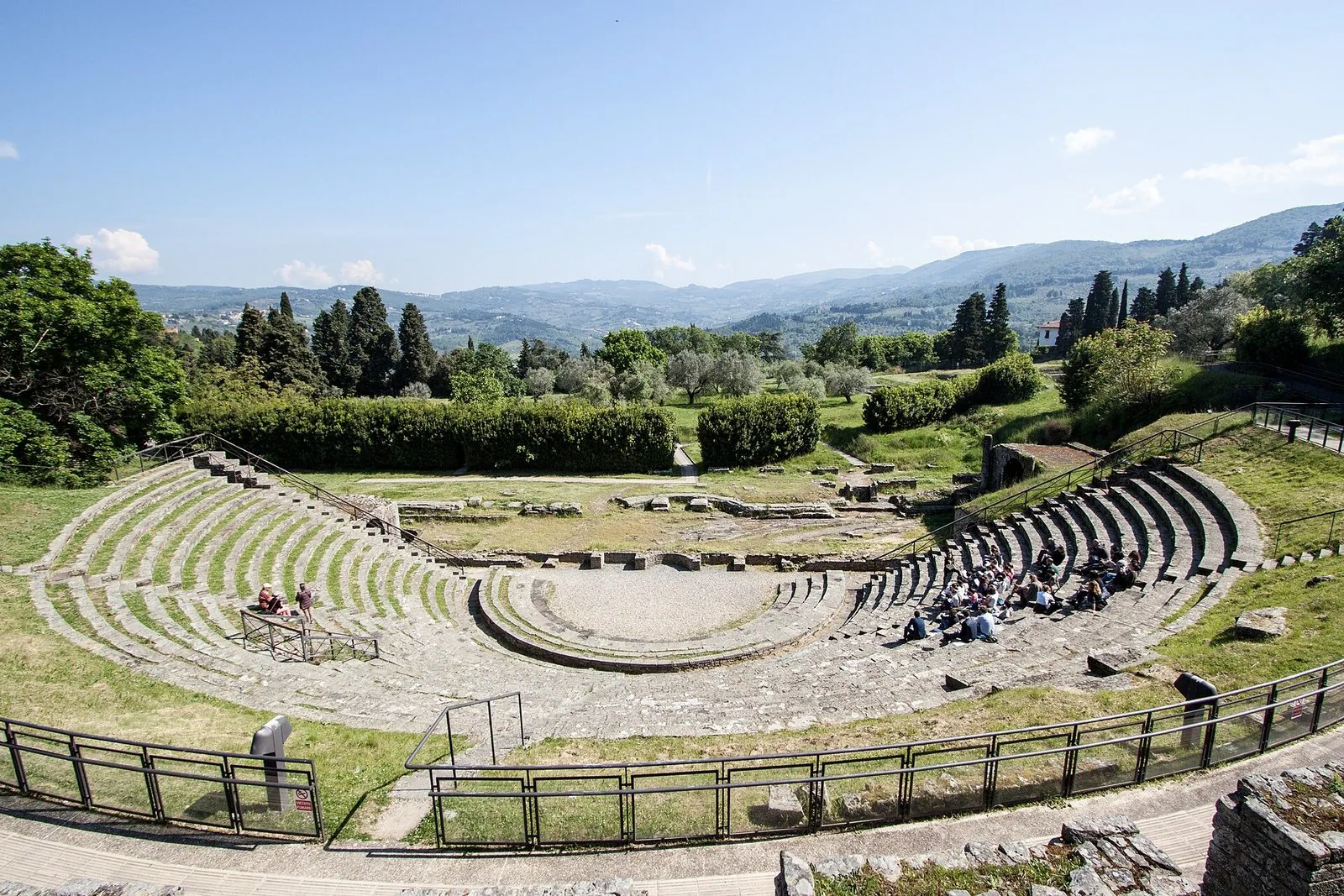 Il teatro romano di Fiesole visto dall'alto in una giornata di sole: la cavea semicircolare di gradini in pietra, l'orchestra, i prati e i cipressi dell'area archeologica e sullo sfondo le colline toscane