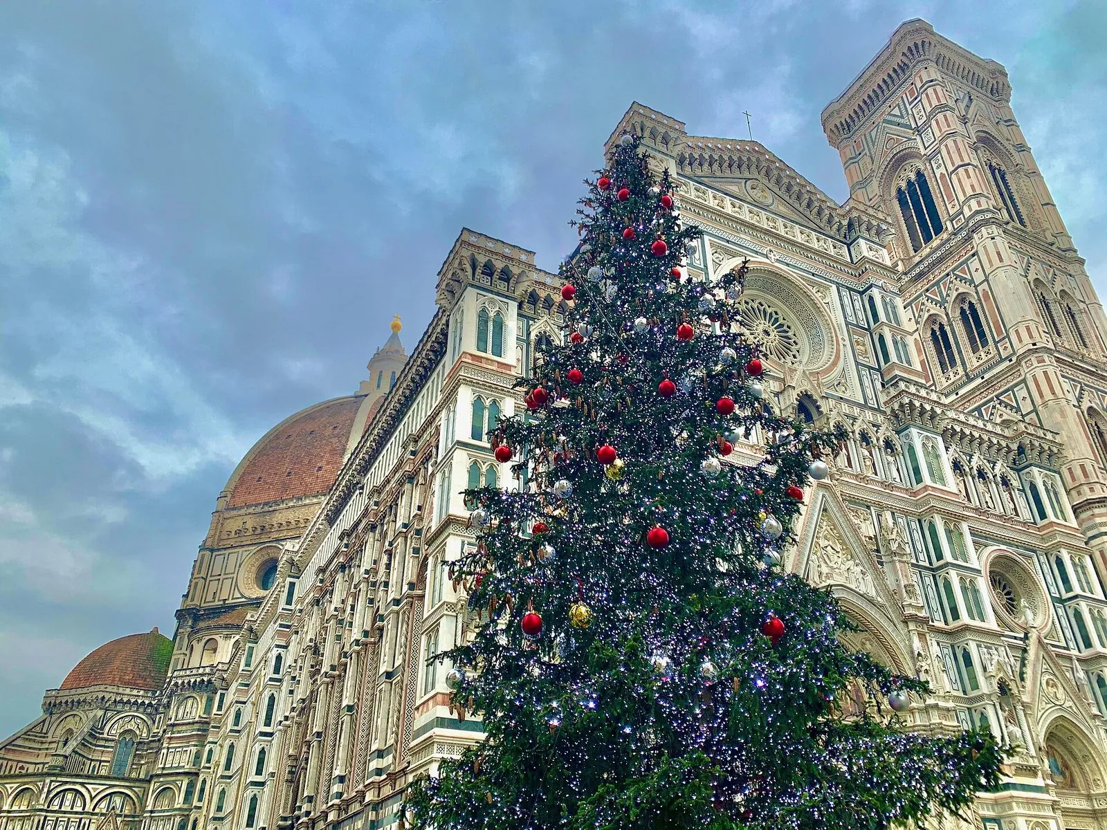 Un grande albero di Natale addobbato con palline rosse, argento e oro e luci bianche davanti alla facciata in marmo bianco, verde e rosa del Duomo di Firenze, con la cupola a sinistra e il Campanile di Giotto a destra sotto un cielo nuvoloso