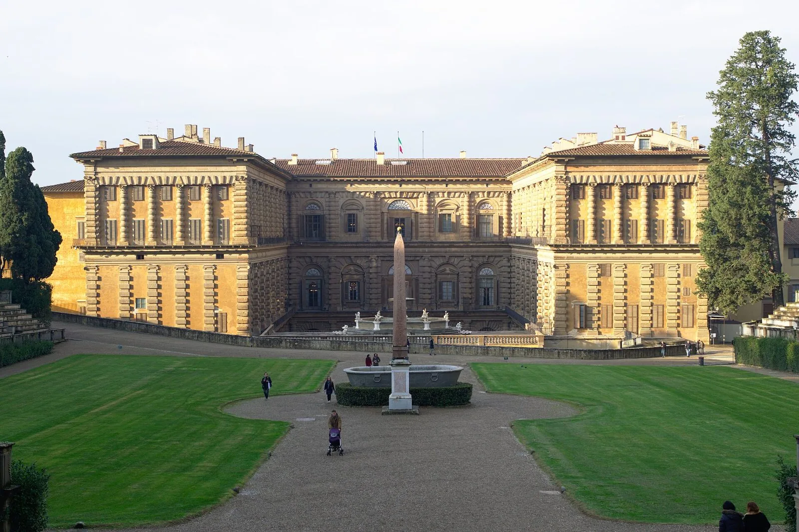 La facciata posteriore di Palazzo Pitti vista dal Giardino di Boboli: il grande palazzo in pietra dorata dal sole, al centro un obelisco accanto a una grande vasca in pietra, prati all'inglese ai lati del viale di ghiaia e alti cipressi