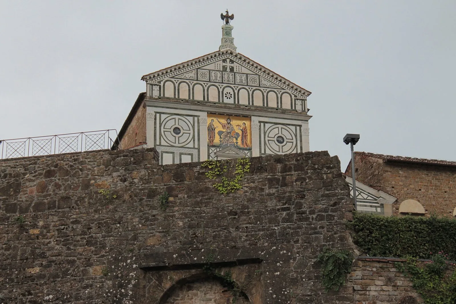 La parte alta della facciata di San Miniato al Monte, in marmo bianco e verde con il mosaico dorato del Cristo tra la Madonna e san Miniato, che spunta sopra un antico muro in pietra sotto un cielo grigio