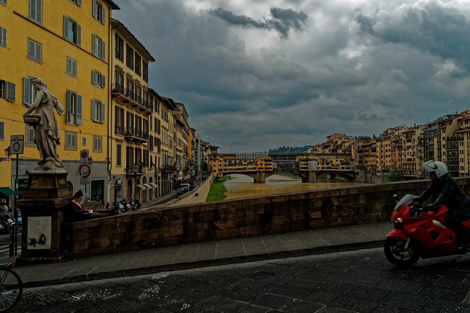 Vista dal Ponte Santa Trinita sotto un cielo grigio e nuvoloso: la statua della Primavera sul parapetto, i palazzi gialli del lungarno e, in fondo, il Ponte Vecchio sull'Arno