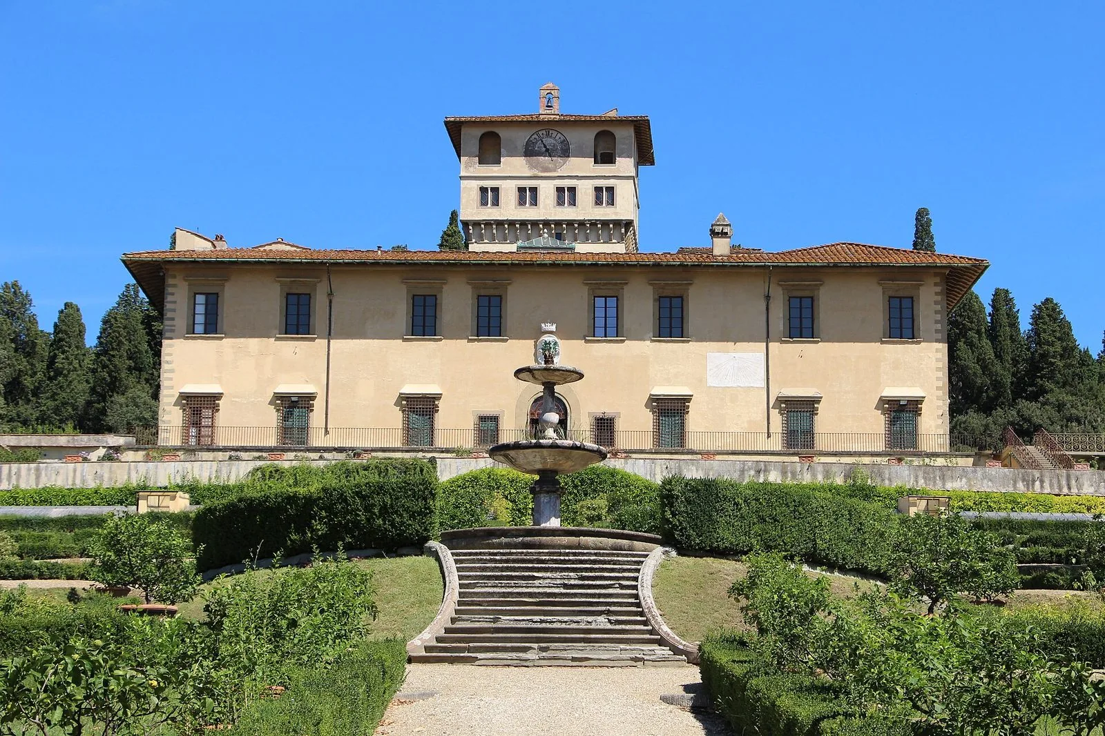 La Villa medicea La Petraia vista di fronte dal giardino: la facciata color sabbia con la torre e l'orologio, una fontana a due vasche e una scalinata in pietra tra siepi e piante di agrumi, sotto un cielo azzurro